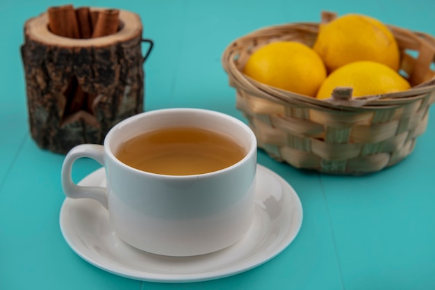 Side view of cup of tea with bowl of cinnamon and basket of lemons on blue background