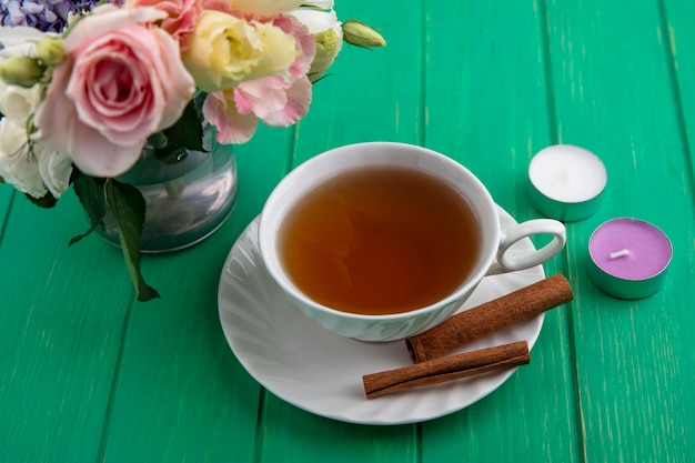 Side view of cup of tea with cinnamon on saucer and candles with flowers on green background