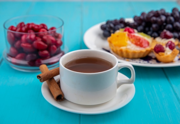 Side view of  a cup of tea with cinnamon sticks with cornel berries on a glass bowl on a blue wooden background