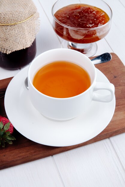 Side view of a cup of tea with strawberry jam in a glass vase on wood board on white