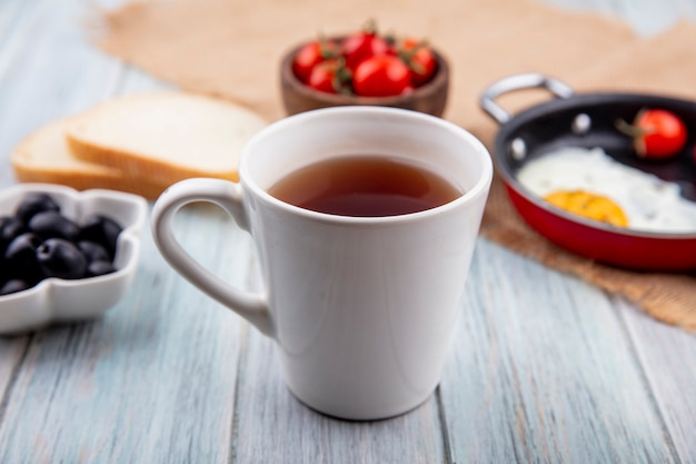 Side view of cup of tea with tomato fried egg on sackcloth and black olive bread slices on wooden surface