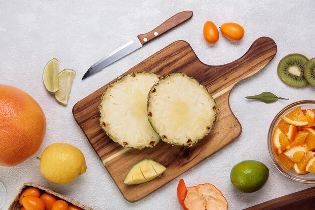 Side view of cut pineapple on cutting board with orange lemon kumquat with knife on white background