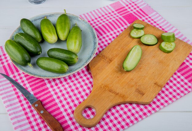 Side view of cut and sliced cucumbers on cutting board with whole ones in plate and knife on plaid cloth and wooden table