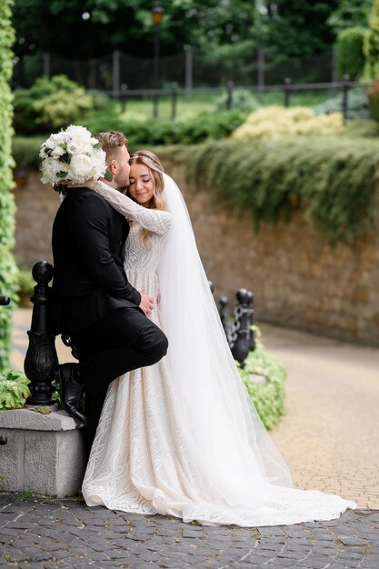 Side view of cute girl dressed in lace gown with dress train embracing her groom and closing eyes during wedding walk in park