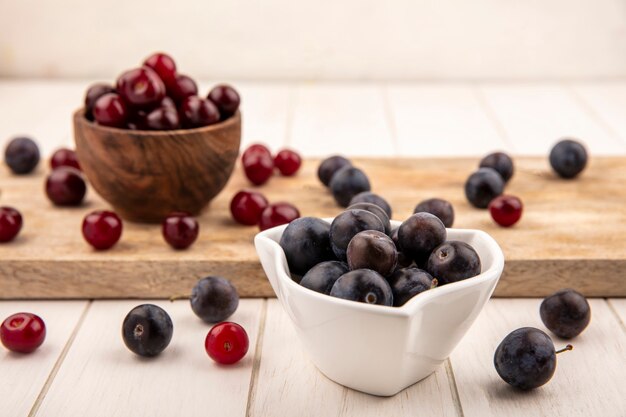 Side view of dark purple sloes on a white bowl with red cherries on a wooden bowl on a wooden kitchen board on a white wooden background