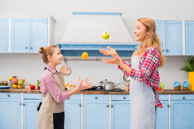 Side view of a daughter and her mother throwing apple and lemon in air at kitchen