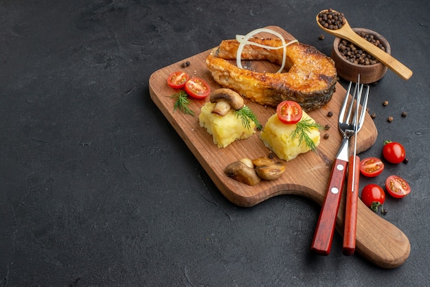 Side view of delicious fried fish and mushrooms tomatoes greens on wooden cutting board cutlery set pepper on black surface