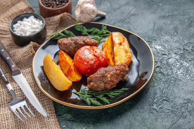 Side view of delicious meat cutlets baked with potatoes and tomatoes on a black plate spices garlics cutlery set on green black mix colors background