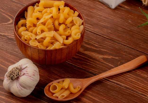 Side view of different macaronis in wooden bowl and spoon with garlic on wooden table