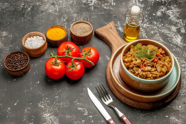 Side view dish tomatoes with penicels plate of the appetizing green beans with tomatoes on the board next to the fork knife bottle of oil and colorful spices on the dark table