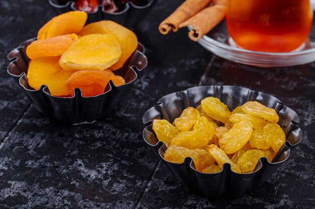 Side view of dried raisins and dried apricots in mini tart tins served with tea on black wooden background