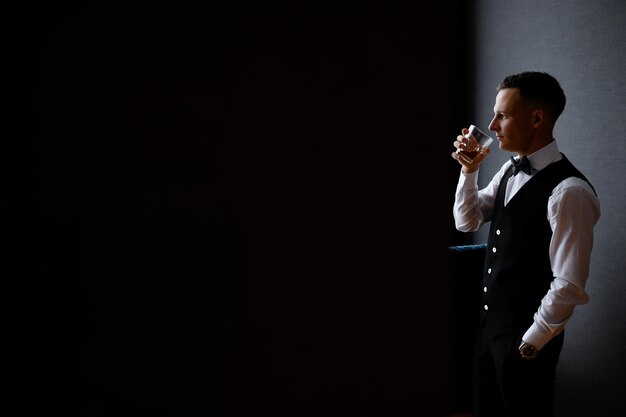 Side view of elegant groom in stylish outfit which standing opposite window keeping hand in pocket drinking alcohol and looking away during wedding morning Concept of wedding and copy space