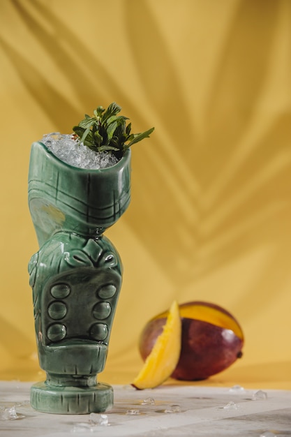 Free photo side view of exotic cocktail in a tiki glass and fresh mango on yellow wall