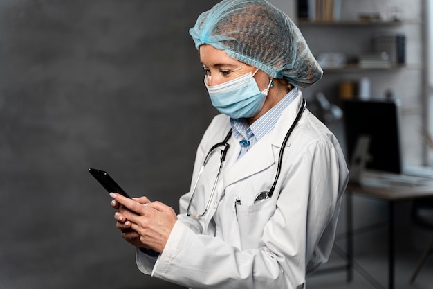 Free photo side view of female doctor with medical mask and hairnet holding smartphone