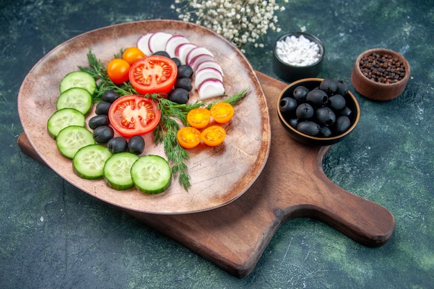 Side view of fresh chopped vegetables in a brown plate on wooden cutting board olives in bowl salt garlics flower on mixed colors background