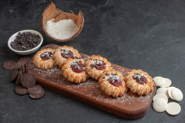 Side view of freshly-baked jam-filled thumbprint cookies on wooden tray and various chocolates on dark surface