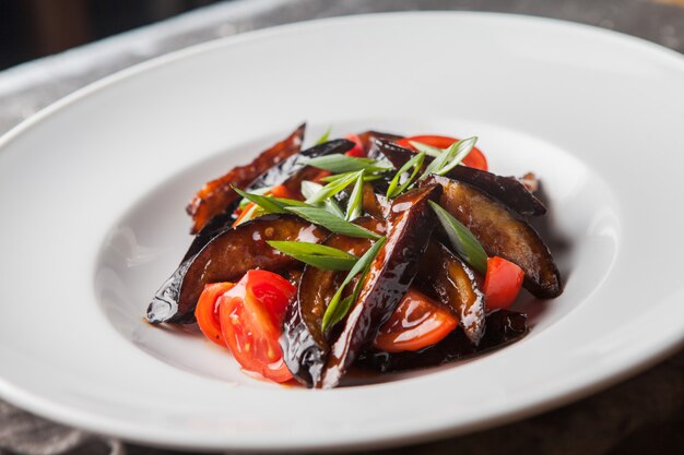 Side view fried eggplant with tomato and chopped green onions and rag in round white plate