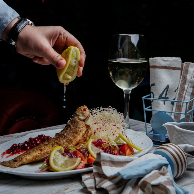 Side view fried fish with glass of wine and lemon and human hand in white plate