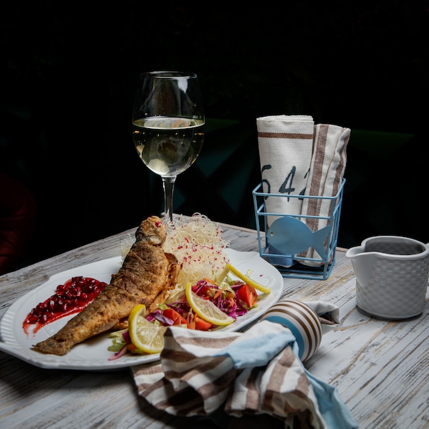 Free photo side view fried fish with glass of wine and lemon and napkins in white plate