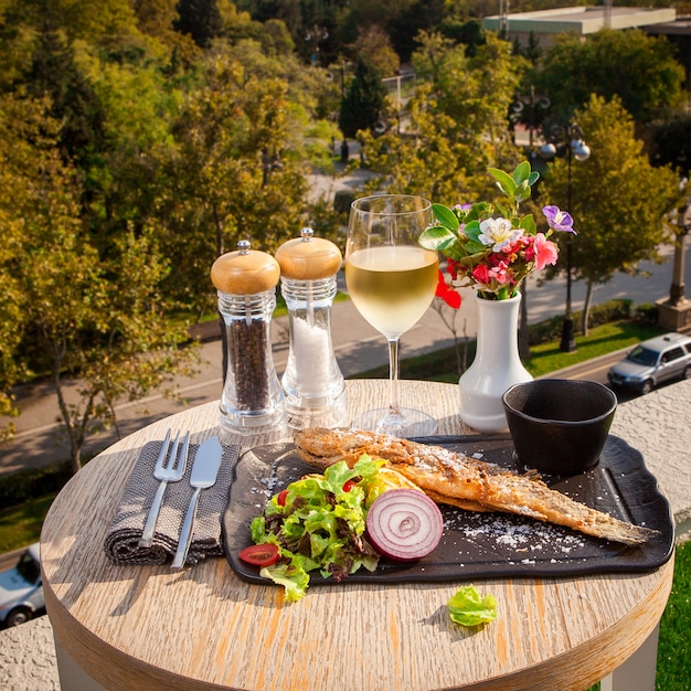 Side view fried fish with lettuce, tomatoes, onions and sauce on a black plate, a glass of white wine on a small round table overlooking the city