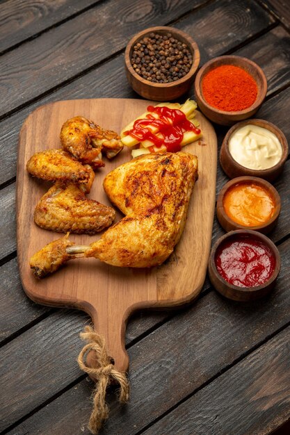 Side view from afar chicken and sauces chicken and french fries on the wooden board next to the black pepper ketchup sauces and spices on the table