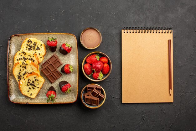 Side view from afar cup of tea with cake plate of cake with chocolate-covered strawberries cup of tea chocolate cream and strawberries in bowls next to pencil and notebook