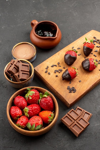 Free Photo side view from afar strawberries on board chocolate cream and strawberries and chocolate-covered strawberries on the kitchen cutting board on the dark table