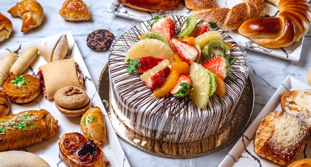 Side view fruit cake with vanilla cream chocolate kiwi orange strawberry pineapple and pastries on the table