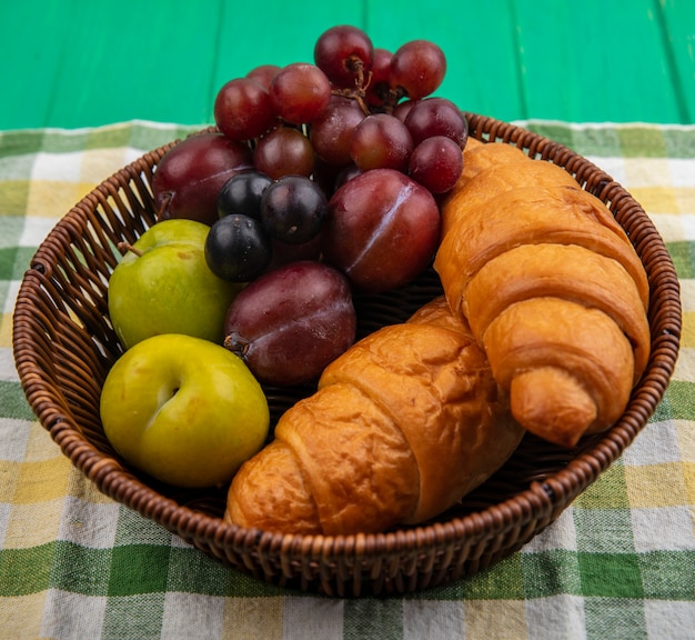 Side view of fruits as grape pluots sloe berries with croissants in basket on plaid cloth and green background