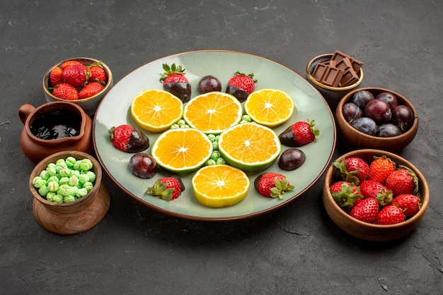 Free photo side view fruits on plate chocolate-covered strawberries and chopped orange in white plate next to the bowls of chocolate sauce sweets and berries on the table