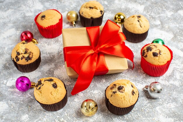 Side view of gift with red ribbon among freshly baked delicious small cupcakes and decoration accessories on ice table