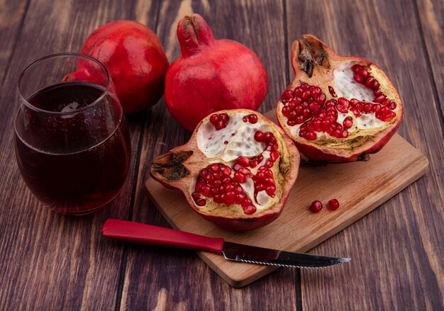 Side view glass of pomegranate juice with pomegranate slices on a cutting board with a knife on a wooden wall