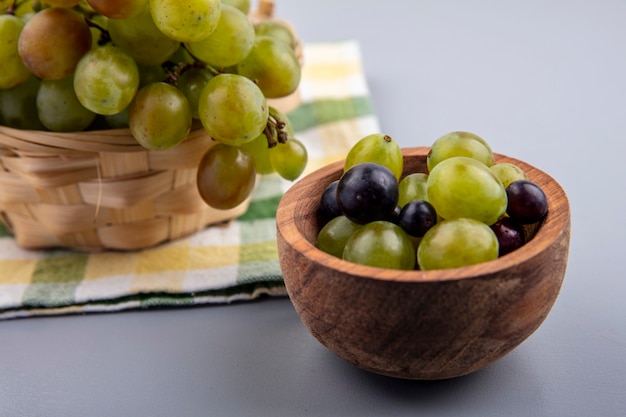 Side view of grape berries in bowl with basket of grape on plaid cloth and gray background