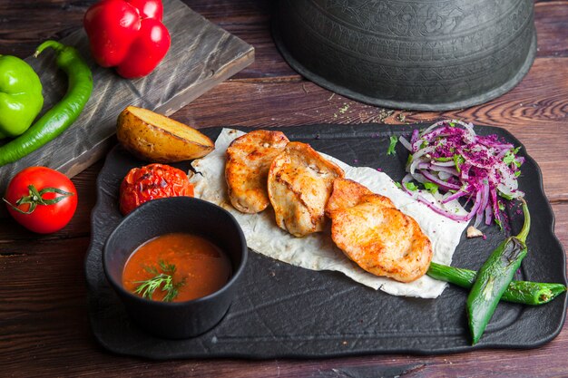 Side view grilled chicken with potatoes, tomato, onion, pepper and sauce on a dark wooden table horizontal