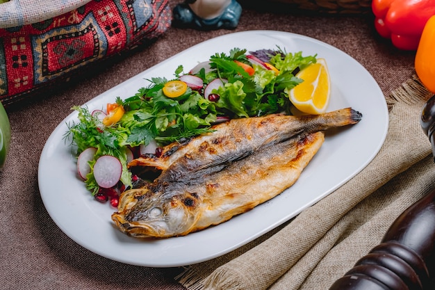 Side view grilled fish with a salad of vegetables and herbs with a slice of lemon