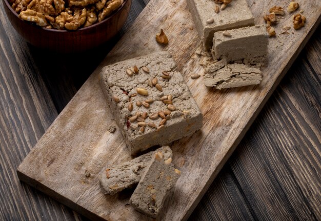 Side view of halva with sunflower seeds on a wooden board and a bowl of walnuts on rustic