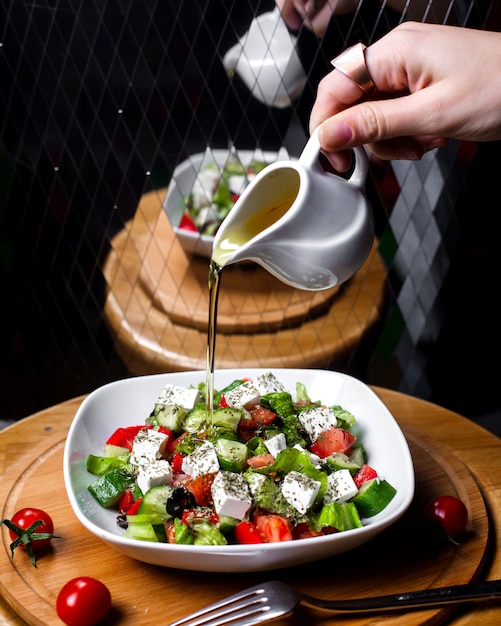 Free photo side view of hand pouring olive oil on fresh salad with feta cheese tomatoe cucumbers in white bowl