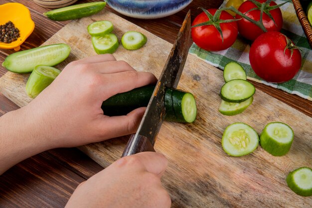 Side view of hands cutting cucumber with knife on cutting board with tomatoes black pepper on wooden surface