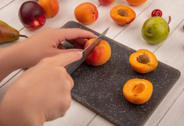 Side view of hands cutting peach with knife and half cut peach on cutting board with pattern of pears peaches on wooden background