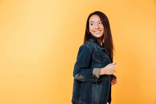 Side view of happy asian woman in denim jacket posing and looking back over yellow background