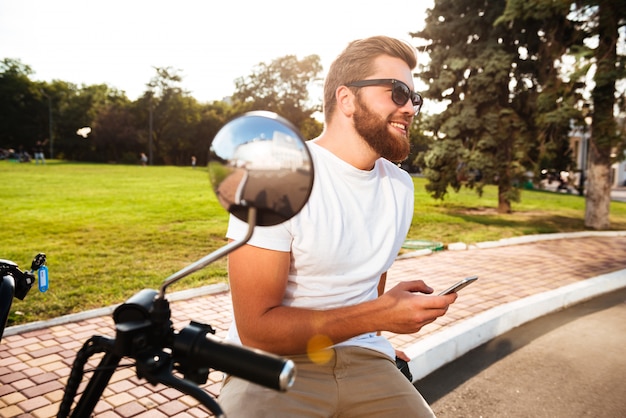 Free photo side view of happy bearded man in sunglasses sitting on modern motorbike outdoors with smartphone and looking away