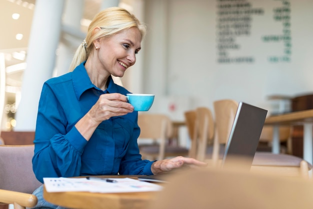 Side view of happy older business woman having cup of coffee and working on laptop