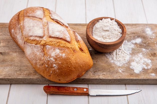 Side view of homemade wheat bread with bowl of flour on cutting board and knife on wooden background