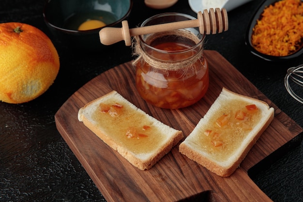 Side view of jar with quince jam and bread slices smeared with quince jam on cutting board and grapefruit grated orange zest on black background