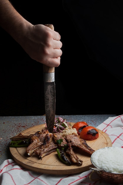 Free photo side view kebab ribs with fried vegetables and chopped onion and human hand and knife and ayran in wooden food tray