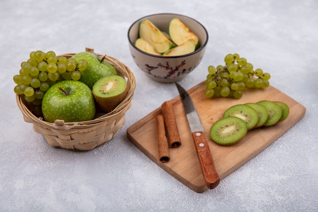 Side view kiwi slices with grapes  cinnamon and a knife on a cutting board with green apples in a basket and slices in a bowl on a white background