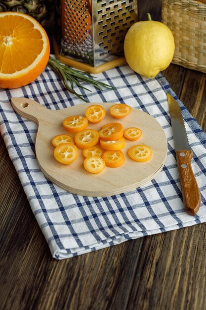 Side view of kumquat slices on cutting board with lemon cut orange scallion grater knife on plaid cloth on wooden background