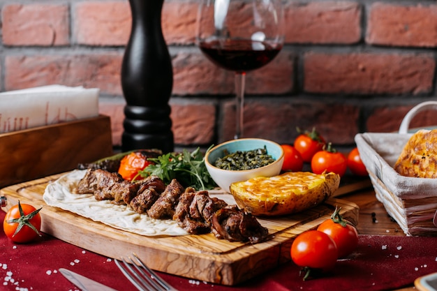 Side view of lamb kebab with baked potato and vegetables on a wooden board