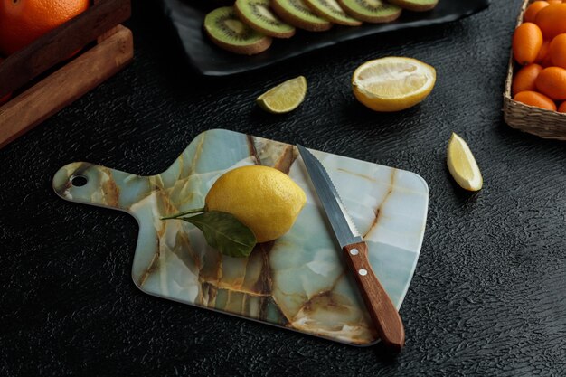 Side view of lemon with knife on cutting board with kiwi slices lime slice cut lemon and whole kumquats on black background