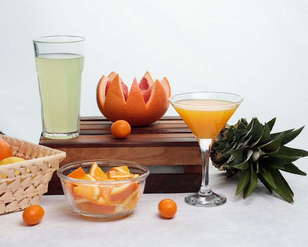 Side view of lime juice in glass and cut grapefruit with kumquat on cutting board with orange slices in bowl pineapple and glass of orange juice on white background